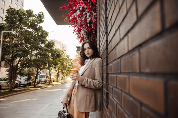 Tall stylish girl with coffee in a beige oversized jacket and orange pants on brick wall background