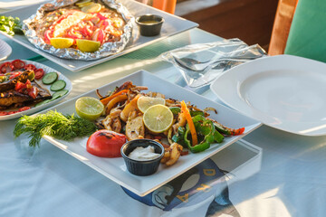 Close-up view of served table in a fish restaurant - grilled squid and vegetables dish, served with dill, lemon and sauce, dishes with grilled fish, seafood and vegetables and one empty plate
