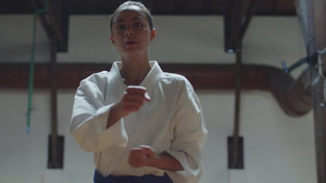 Front view of girl in kimono kicking and punching at camera. Medium shot of focused and beautiful Caucasian teenager practicing karate, rehearsing movements for competition.Martial arts, sport concept