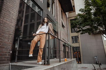  Tall stylish girl with oversized jacket posing near the railing and steps on the city background