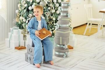 baby boy in blue pajamas sitting on boxes with Christmas gifts on the background of a Christmas tree and a pile of gifts.