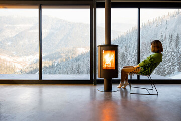 Woman sitting near fireplace at modern living room with great view on snowy mountains. Concept of...