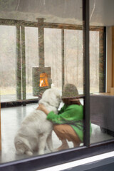 Young woman with her dog at modern house on nature. Girl in green sweater sitting near the window and look on fireplace. Rear view through the window, focus on the fireplace