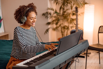 Joyful multiracial woman in headphones sitting on bed and smiling while playing melody on...