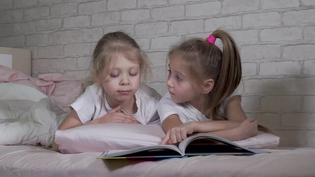 Children read a book, look at pictures, study while lying in bed. Girls in the bedroom. Two preschooler children. Beautiful, funny, cute, smart kids
