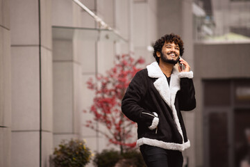 Positive indian man talking on smartphone and looking up on urban street 