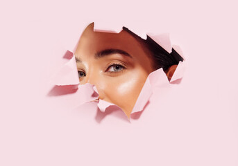 Face of a young beautiful woman with clean skin and natural makeup, peeking into a hole in pink paper