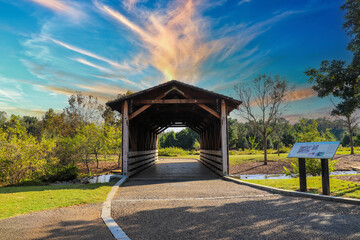 a shot of a wooden covered bridge over a river surrounded by lush green and autumn colored trees with lush green grass, blue sky and clouds at sunset at Garrard Landing Park in Alpharetta Georgia USA	 - obrazy, fototapety, plakaty