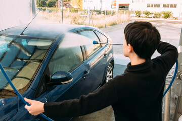 Close-up image of a woman cleaning a blue car using a hose with high pressure water. Concept of self-service car washing. 