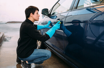 Young woman cleaning the door of a blue car using a sponge wearing blue rubber gloves. Concept of self-service car washing. 