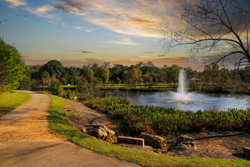 a shot of winding footpath along a lake with a water fountain surrounded by lush green and autumn colored trees and plants with powerful clouds at sunset at Garrard Landing Park in Alpharetta Georgia - obrazy, fototapety, plakaty