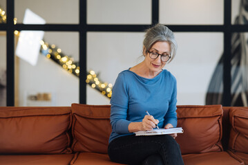 Happy senior woman in glasses writing to notebook at home