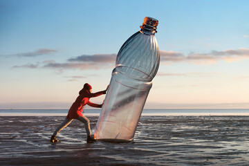 A woman is pushing a huge plastic bottle on the shore. Environmental pollution by plastic. Recyclable packaging.