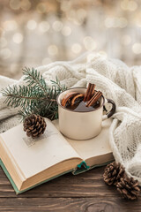 Obraz na płótnie Canvas Mug of warming drink with cinnamon sticks on a book on a background of fir branches and cones. Knitted white scarf envelops a mug of hot mulled wine with spices on a wooden background. Cozy winter