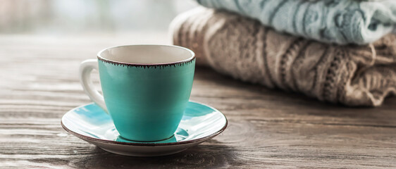 A cup of coffee on the background of a stack of cozy winter autumn sweaters in pastel colors on a wooden background. Knitted woolen sweaters. Fashion Style. Copy space.Banner	