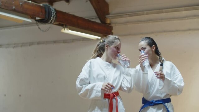 Front view of martial artsists drinking water and talking in gym. Medium shot of cheerful and smiling Caucasian girls in kimonos recovering strength after karate workout. Sport, friendship concept