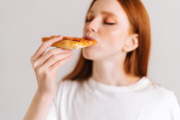 Close-up face of happy attractive young woman with closed eyes appetite eating delicious pizza standing on white isolated background. Pretty redhead female eating tasty meal, selective focus.