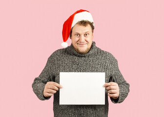 A man in a gray sweater and a red New Year's Eve is holding an empty white sheet of paper in his hands. New Year's mood. Template for text, mockup.