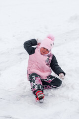 Child is playing snowballs. Happy little girl in warm suit is sitting on snowy road and enjoying rest in nature. 