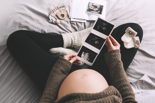 Pregnant woman with ultrasound image and medical test reports in cozy knitted sweater in bed at cold winter days. Expectant mother with pregnant belly waiting for baby. Concept of pregnancy.