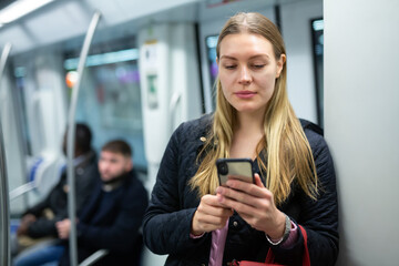 Positive young woman absorbed in her smartphone while traveling in subway car
