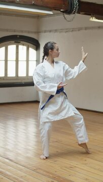 Vertical shot of serious girl performing karate pose in gym. Long shot of confident female martial artsist wearing kimono with blue belt and standing in self defence pose in gym. Sport concept