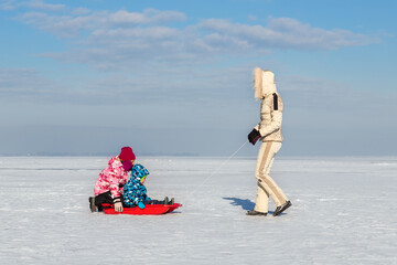 Fototapeta na wymiar Young adult mother wear warm jacket enjoy have fun sledging two cute little sibling kids boy girl at frozen white snow lake field against clean blue sky landscape bright sunny cold winter day. Outdoor
