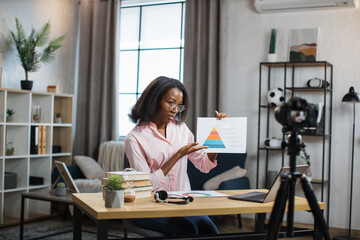 Pleasant young woman showing financial charts while recording video on modern camera. African american freelancer working remotely while staying at home.