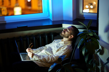 Exhausted young businessman sit with closed eyes in dark office with laptop tired of overtime. Pr...