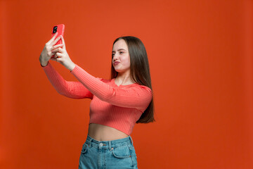 Beautiful modern young girl with long hair hold mobile phone, posing for selfie, looking at camera on orange background