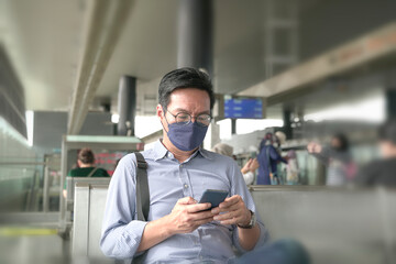 Asian businessman with face mask on looking at his smartphone, waiting on train platform. Masked transit concept.