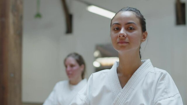 Close-up shot of karateka smiling at camera while sitting in gym. Front view of cheerful martial artsist in white kimono posing for camera, sitting on floor, enjoying her hobby. Sport, emotion concept