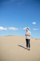 Young tired man standing with bottle water while sitting on the top of sand dune in desert, sand is al around and some greens. Hot summer weather, concept of travelling around planet. Europen desert