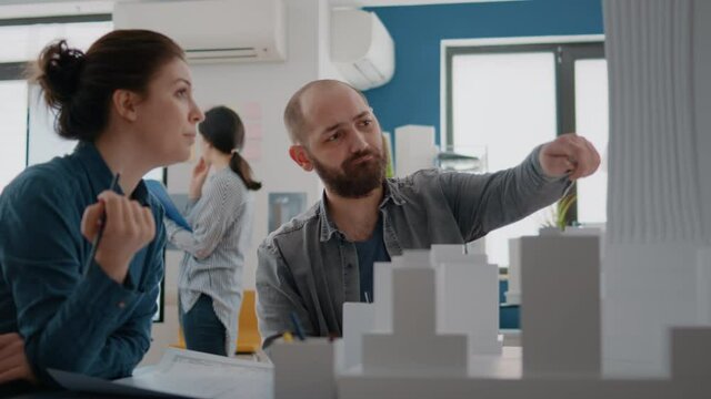 Man and woman pointing at building model to plan blueprints on paper. Colleagues using maquette to design constraction structure, helping with architectural development. Industrial project