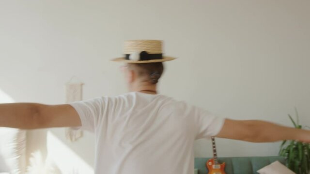 Young fashion creative hipster wearing hat, pink sunglasses guy dancing and rotating around with his old retro fim camera with strap his neck in contemporary inerior. Millennial generation lifestyle
