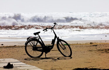 Fototapeta premium Abandoned bicycle on the sand, with slippers, strong sea bottom and big waves. Loneliness, depression, post covid.