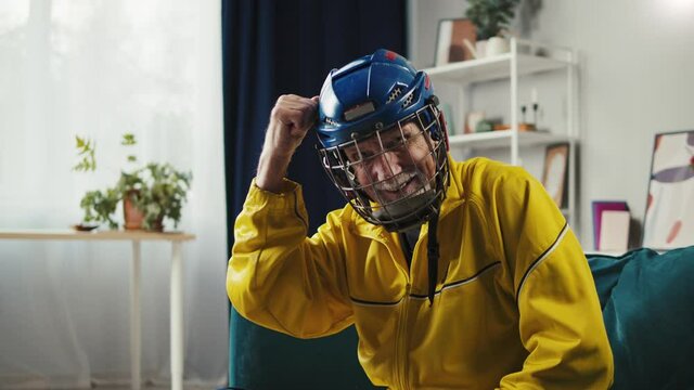 Old funny hockey player portrait. Elderly man wearing sports clothes and helmet. Grey-haired grandfather posing at home, looking in camera. Healthy lifestyle and long life. 
