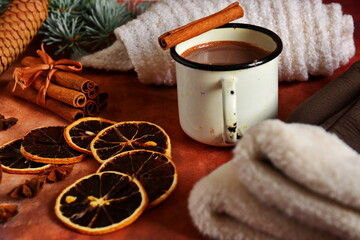 Winter warming drink hot chocolate and cinnamon sticks in tin bowl, woman gloves and white scarf on table decorated with spices, star anise, slice of dry lemon and branch of christmas tree and cones
