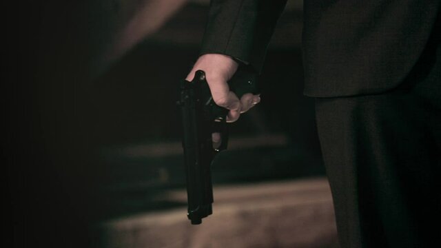 Mysterious mafia agent man in black suit holding pistol by torso. Pan out from shadow