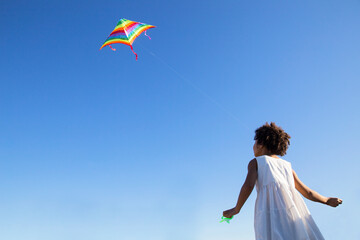 Cute curly, mixed race girl flying a colorful kite in the blue sky.