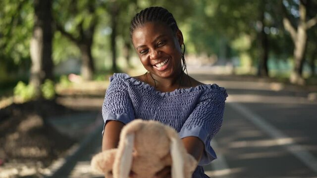Happy millennial African American woman admiring toy rabbit standing in sunny park outdoors. Portrait of positive beautiful plus-size lady enjoying summer weekend in slow motion