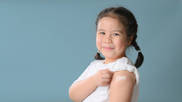 Coronavirus Vaccination Advertisement. Happy Vaccinated Little asian girl Showing Arm With Plaster Bandage After Covid-19 Vaccine Injection Posing Over Blue Background, Smiling To Camera. Blank Space
