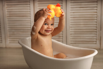 a baby of 11 months is bathing in a white baby bath with rubber ducklings, the baby is laughing,...