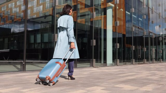 Young businesswoman walking along city street and holding suitcase in hand on autumn day spbi. Back view of beautiful woman walks forward and looks with smile, moves luggage and goes along glass
