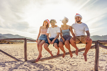 Suggestive photography with backlight sun effect of four young happy people enjoying outdoor life in exotic vacation resort sitting on wooden fence on a white sand beach at sunset - Powered by Adobe