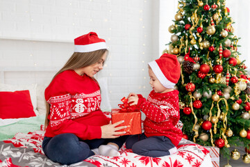 Obraz na płótnie Canvas a pregnant woman with a baby boy in a red sweater and hats give gifts under the Christmas tree at home on the bed and rejoice in the new year and Christmas