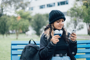 Latin teenager college girl sitting on a bench at the park buying online with credit card