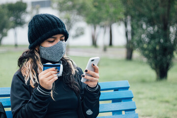 Latin teenager college girl sitting on a bench at the park buying online with credit card and face mask on