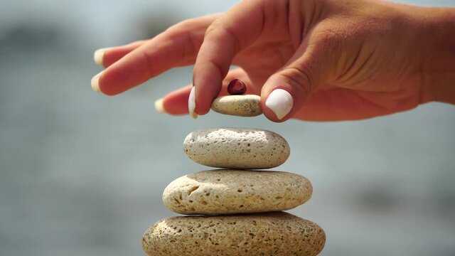 Woman bilds stones pyramid on the seashore on a sunny day on the blue sea background. Happy holidays. Pebble beach, calm sea, travel destination. Concept of happy vacation on the sea, meditation, spa