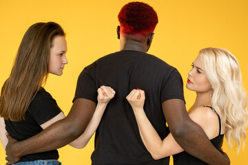 Fototapeta premium Love triangle. Relationship jealousy. Female competition. Multiracial group of African man embracing two fist threatening angry Caucasian women isolated on orange background.